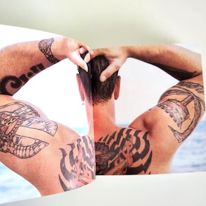 May include: A printed photograph showing the back of a person with short dark hair and multiple black ink tattoos on their shoulders, upper arms, and back. The person has their hands raised to their head, with a blurred blue water background.