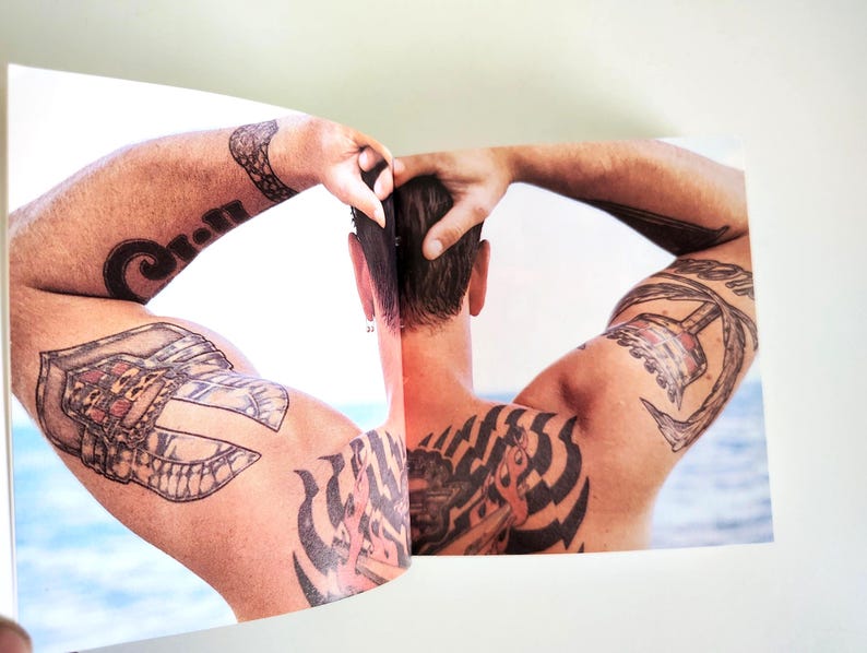 May include: A printed photograph showing the back of a person with short dark hair and multiple black ink tattoos on their shoulders, upper arms, and back. The person has their hands raised to their head, with a blurred blue water background.