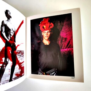 May include: An open book page 117 featuring two photographs. The left side shows a shirtless figure in black shorts with a large red X overlay. The right side shows a figure wearing a black long-sleeved shirt and a bright red headpiece.