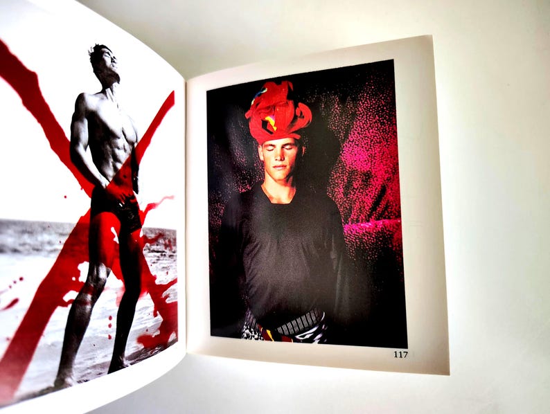 May include: An open book page 117 featuring two photographs. The left side shows a shirtless figure in black shorts with a large red X overlay. The right side shows a figure wearing a black long-sleeved shirt and a bright red headpiece.