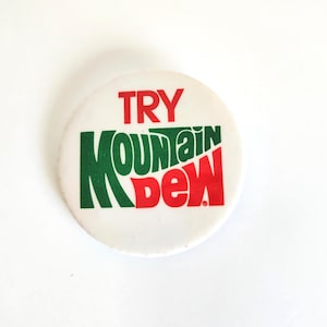 May include: A white circular button with the text "TRY MOUNTAIN DEW" in red and green. The button has a retro design, with the words stacked on top of each other. The button is likely a promotional item for the soft drink.