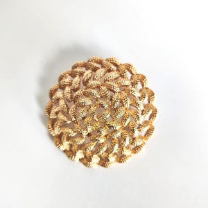 May include: A gold-toned, round brooch with a textured, woven design. The brooch is made of a shiny, metallic material and is set against a white background. The intricate pattern gives the brooch a vintage or antique appearance.