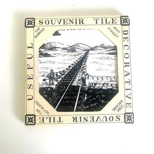 May include: Souvenir tile with a black and white landscape scene of a railway track and figures. The tile is heat proof with a cork backing. The packaging reads "Souvenir Tile" and "Useful for Decorative".