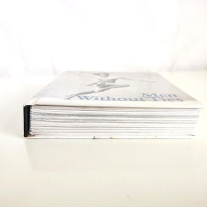 May include: A hardcover book titled Men Without Ties with a white cover featuring a black and white illustration of a figure in motion. The book is positioned horizontally, showing the white paper edges of the pages and a black spine detail.