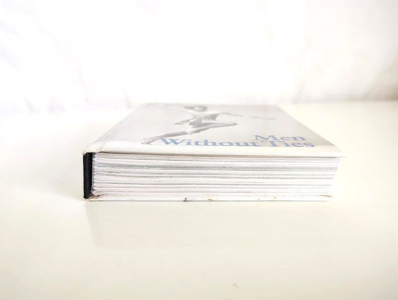 May include: A hardcover book titled Men Without Ties with a white cover featuring a black and white illustration of a figure in motion. The book is positioned horizontally, showing the white paper edges of the pages and a black spine detail.