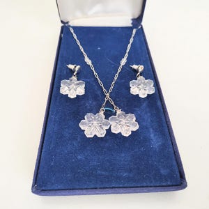May include: A jewelry set featuring a necklace and earrings, presented in a blue velvet box. The necklace and earrings have a snowflake design. The jewelry is silver-toned with clear, faceted crystal snowflake pendants and matching earrings.