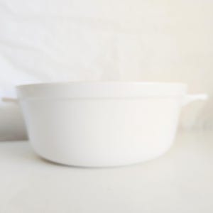 May include: A white ceramic casserole dish with two small handles. The dish has a rounded shape and is sitting on a white surface. The background is a white, slightly textured fabric.