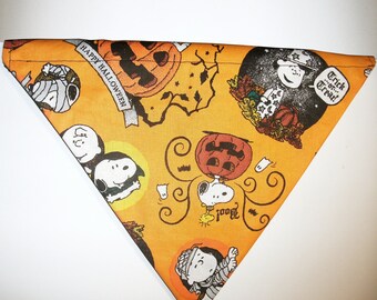 May include: Orange triangular bandana with a Halloween theme featuring Peanuts characters like Snoopy and Woodstock. The bandana has a black and white illustration of the characters dressed in Halloween costumes. The bandana has the text "Happy Halloween" and "Trick or Treat!" printed on it.