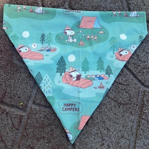 Puede incluir: Bandana triangular para mascotas con temática de camping. La tela turquesa presenta a Snoopy con sombrero de vaquero, tiendas de campaña, árboles y las palabras "CAMPERS" y "HAPPY CAMPERS". El diseño es un patrón repetitivo.