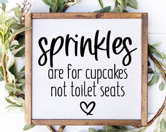 May include: A wooden sign with the text "sprinkles are for cupcakes not toilet seats" in black lettering. A small heart is below the text. The sign is surrounded by green leafy branches.