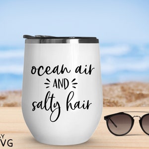 May include: White stainless steel tumbler with black text that reads "ocean air and salty hair". The tumbler is sitting on a wooden surface with a pair of sunglasses next to it.
