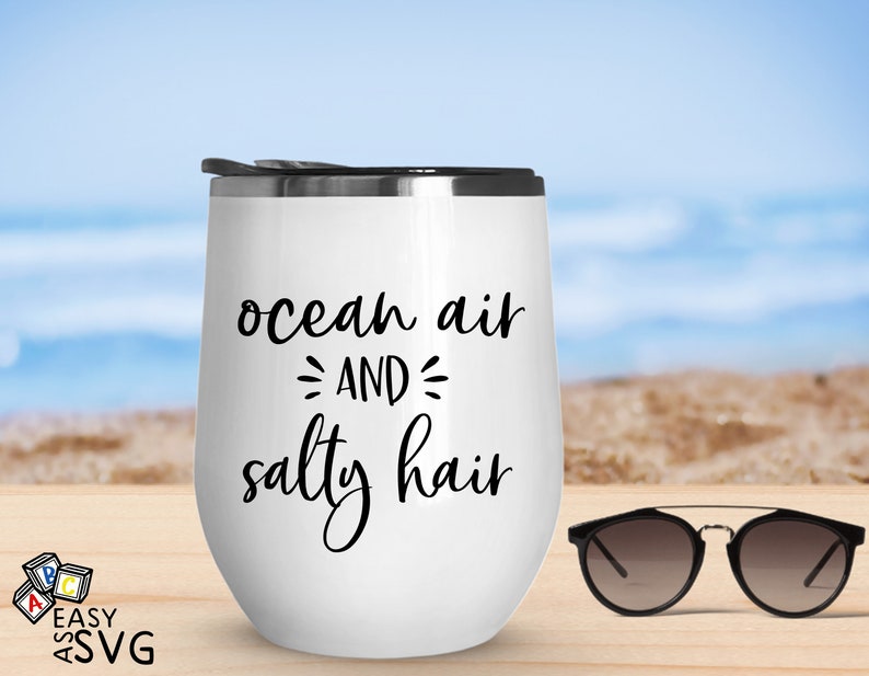May include: White stainless steel tumbler with black text that reads "ocean air and salty hair". The tumbler is sitting on a wooden surface with a pair of sunglasses next to it.