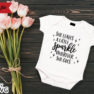 May include: White baby onesie with black text that reads "She leaves a little sparkle wherever she goes". The onesie is on a dark wood background with a bouquet of pink tulips.