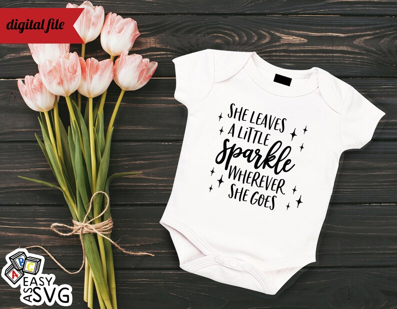 May include: White baby onesie with black text that reads "She leaves a little sparkle wherever she goes". The onesie is on a dark wood background with a bouquet of pink tulips.