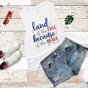 May include: White tank top with the text "land of the free because of the brave" in blue, red, and gray lettering. The tank top is laying on a white wooden surface with a pair of white sneakers, red sunglasses, and a pair of blue denim shorts.