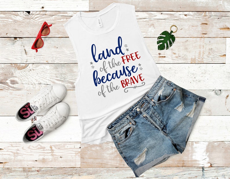 May include: White tank top with the text "land of the free because of the brave" in blue, red, and gray lettering. The tank top is laying on a white wooden surface with a pair of white sneakers, red sunglasses, and a pair of blue denim shorts.