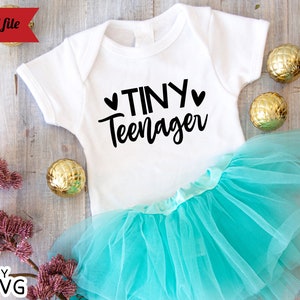 May include: White baby onesie with black text that reads "Tiny Teenager" with two small hearts on either side of the word "Tiny". The onesie is on top of a turquoise tulle tutu.