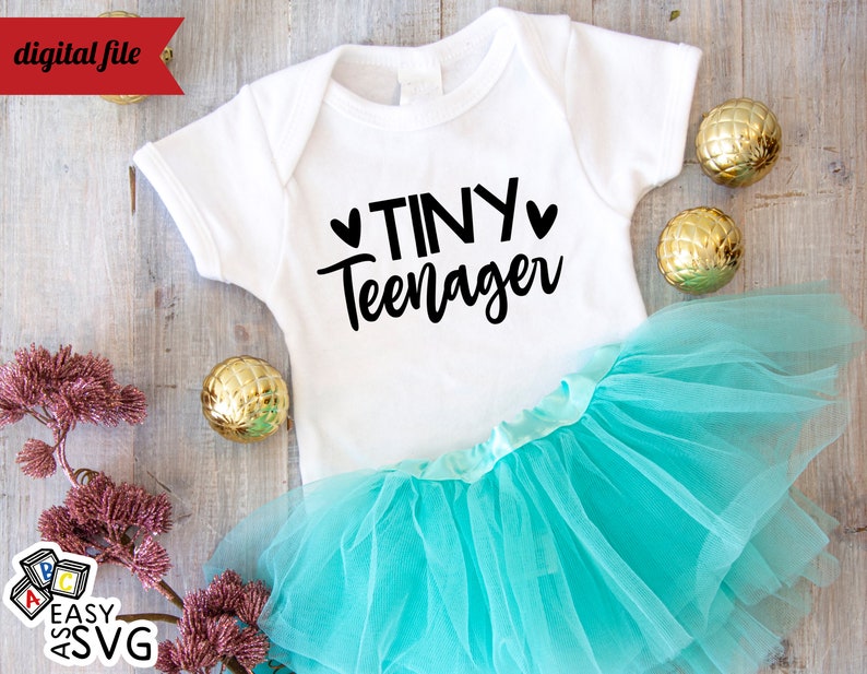 May include: White baby onesie with black text that reads "Tiny Teenager" with two small hearts on either side of the word "Tiny". The onesie is on top of a turquoise tulle tutu.