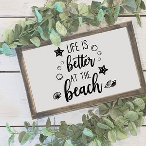 Peut inclure: Un panneau encadr&eacute; avec le texte "LIFE IS better AT THE beach" en &eacute;criture noire et des graphiques d&eacute;coratifs d'&eacute;toiles de mer, de bulles et de coquillages. Le cadre est de couleur marron fonc&eacute; et le fond est blanc. De la verdure entoure le panneau.