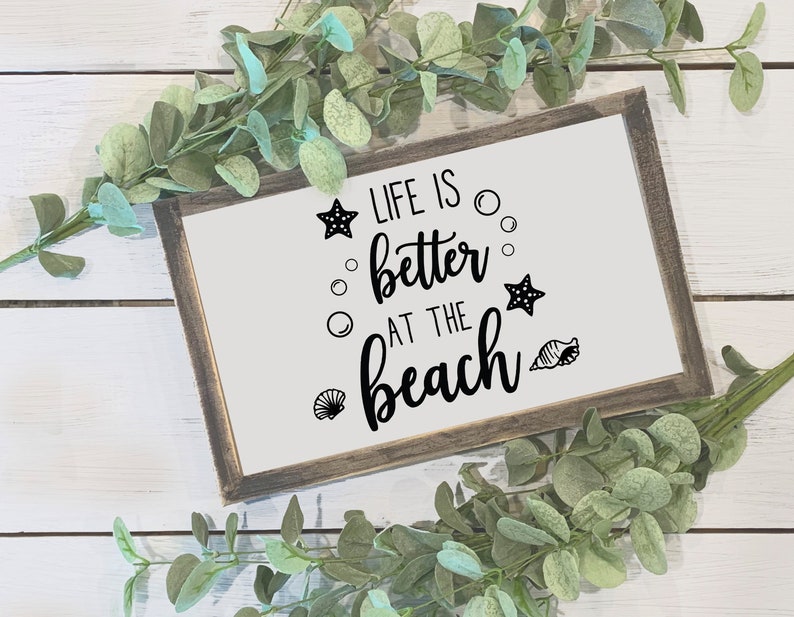 Peut inclure: Un panneau encadr&eacute; avec le texte "LIFE IS better AT THE beach" en &eacute;criture noire et des graphiques d&eacute;coratifs d'&eacute;toiles de mer, de bulles et de coquillages. Le cadre est de couleur marron fonc&eacute; et le fond est blanc. De la verdure entoure le panneau.