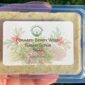 May include: A clear plastic container with a green and brown sugar scrub soap bar inside. The label reads "Foraged Berry Wood Sugar Scrub Soap" and "4 oz/120 ml".