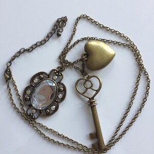 May include: A gold chain necklace with three charms: a large crystal, a heart, and a key. The crystal is surrounded by a decorative metal frame.