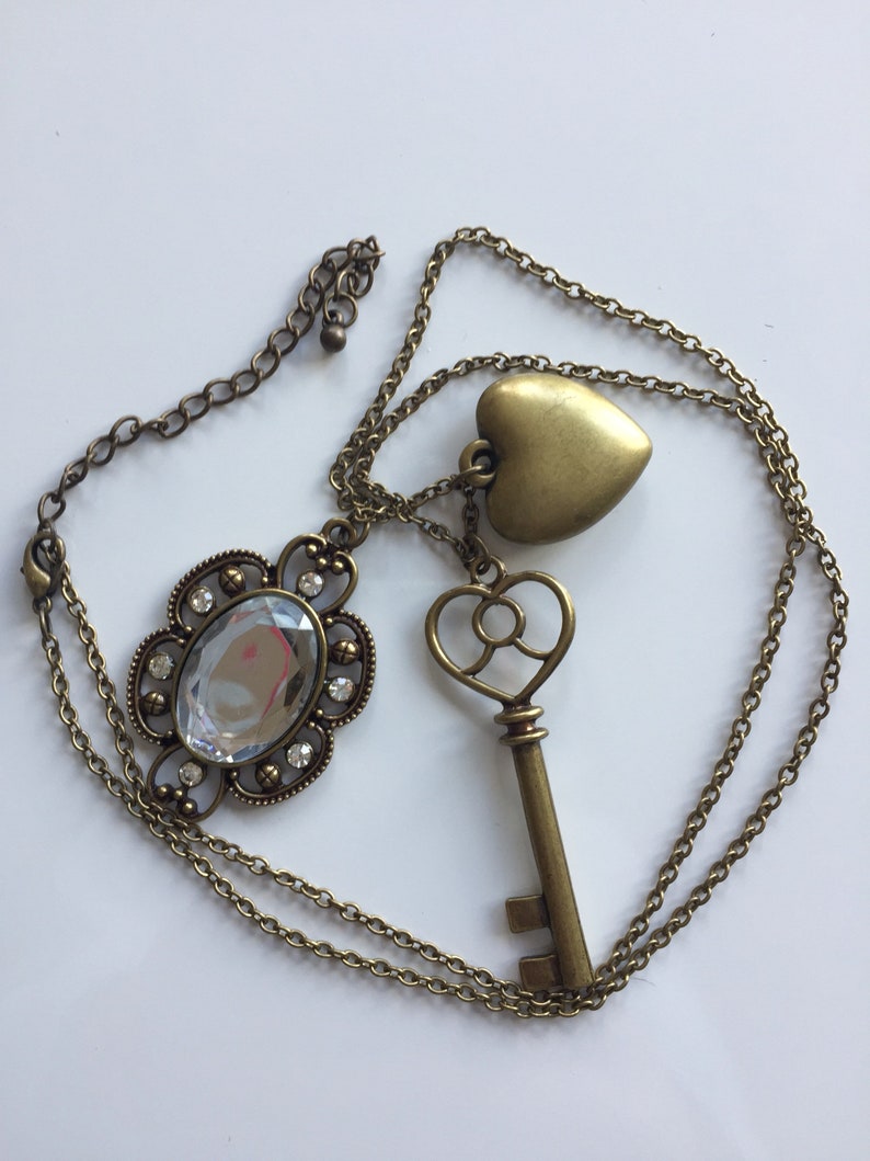 May include: A gold chain necklace with three charms: a large crystal, a heart, and a key. The crystal is surrounded by a decorative metal frame.