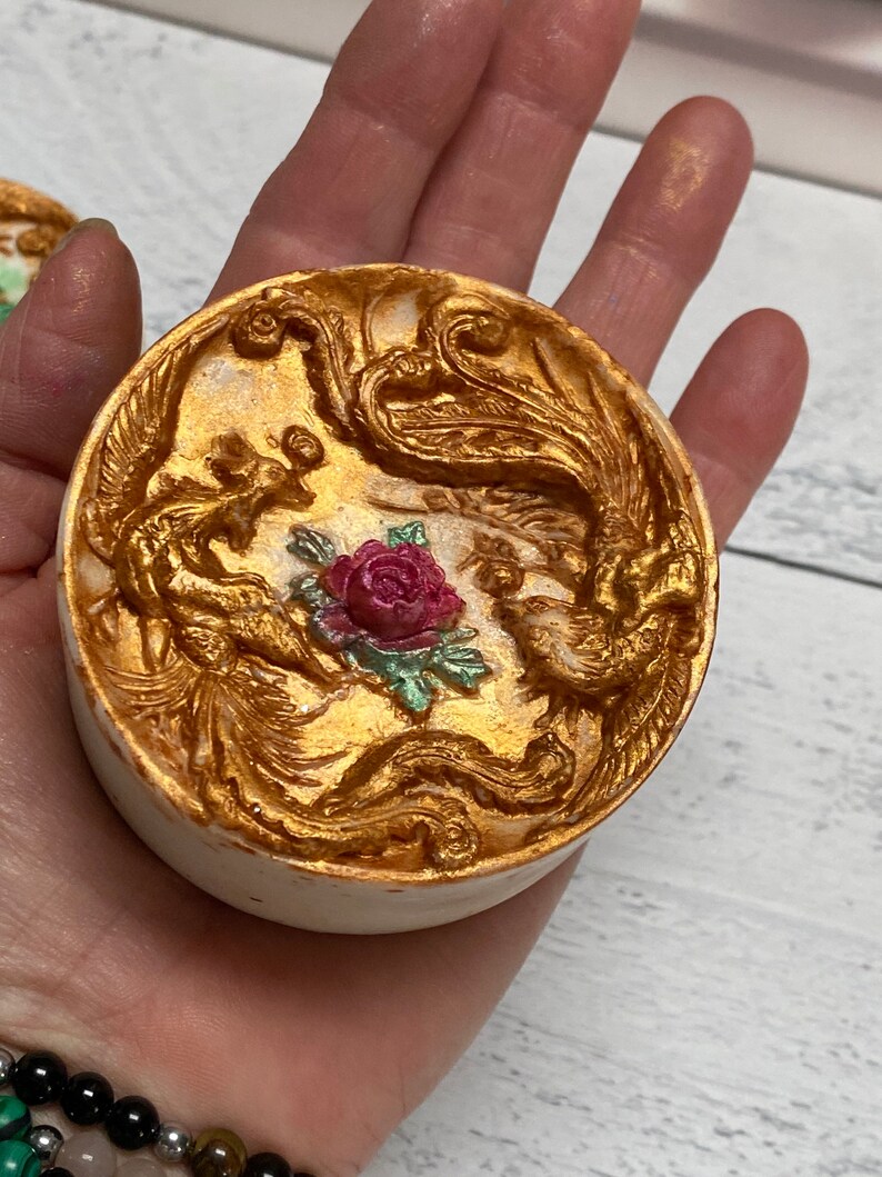 Może przedstawiać: Biała bomba do kąpieli z złotym i czerwonym wzorem. Wz&oacute;r przedstawia dwa stylizowane feniksy z rozpostartymi skrzydłami i czerwoną r&oacute;żę w środku.