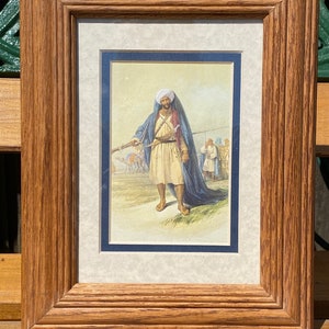 May include: A framed print of a man in traditional clothing holding a rifle. He is wearing a blue turban and a long blue robe. The print is set against a light brown background.