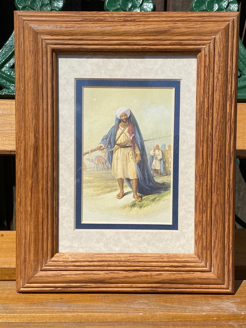 May include: A framed print of a man in traditional clothing holding a rifle. He is wearing a blue turban and a long blue robe. The print is set against a light brown background.