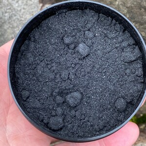 May include: A black plastic container filled with black charcoal powder. The powder is fine and has a slightly grainy texture.