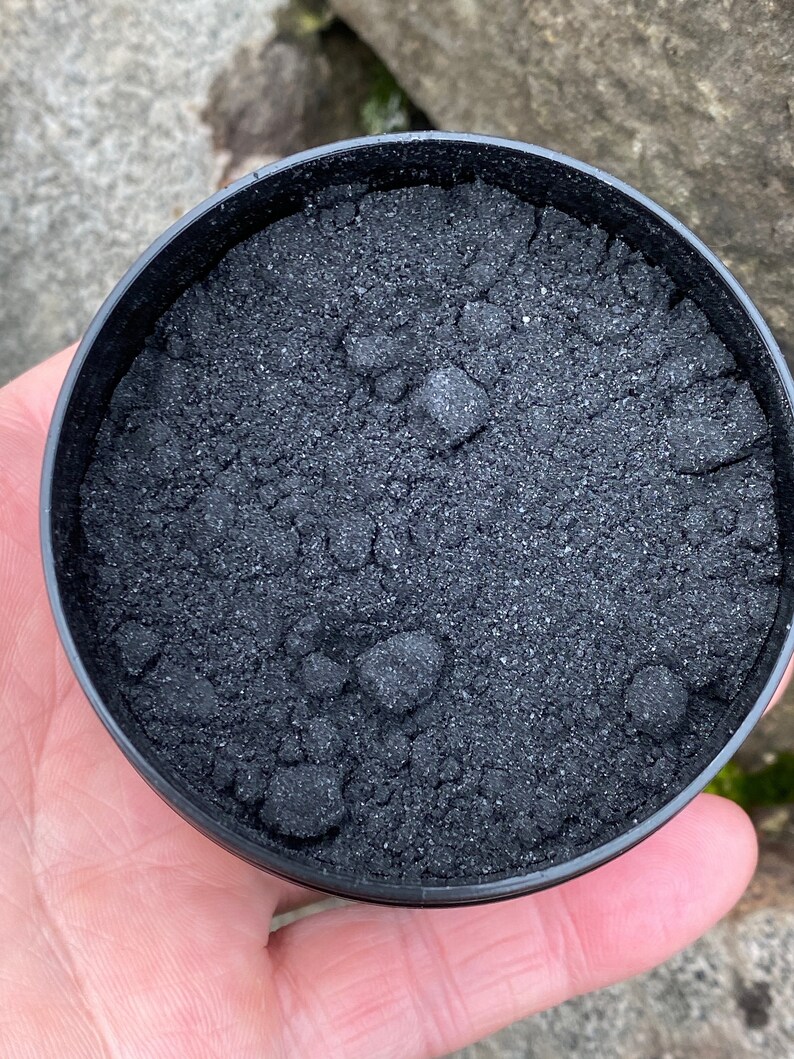 May include: A black plastic container filled with black charcoal powder. The powder is fine and has a slightly grainy texture.