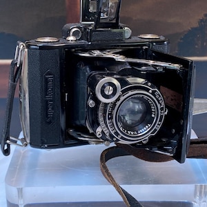 May include: A black Super Ikonta folding camera with a Zeiss Ikon Tessar 1:3.5 lens. The camera features a metal bellows mechanism, a top-mounted viewfinder, and a leather strap. The lens barrel displays technical markings including aperture and shutter speed settings, with the serial number 1672216 clearly visible on the front ring.