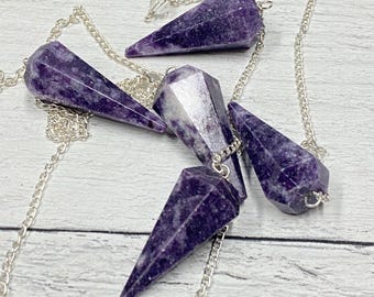 May include: Five purple crystal pendulums with silver chains and a clear crystal bead. The pendulums are faceted and cone-shaped, with varying shades of purple. The chains are linked and attached to the top of each crystal.