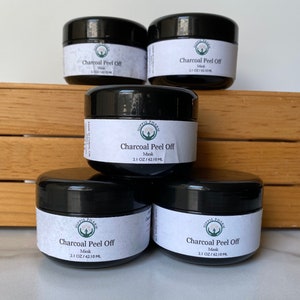 May include: Four black jars of charcoal peel off face mask, each labeled "Hippie Pharm Charcoal Peel Off Mask 2.1 OZ/62.10 ML" on a wooden surface.