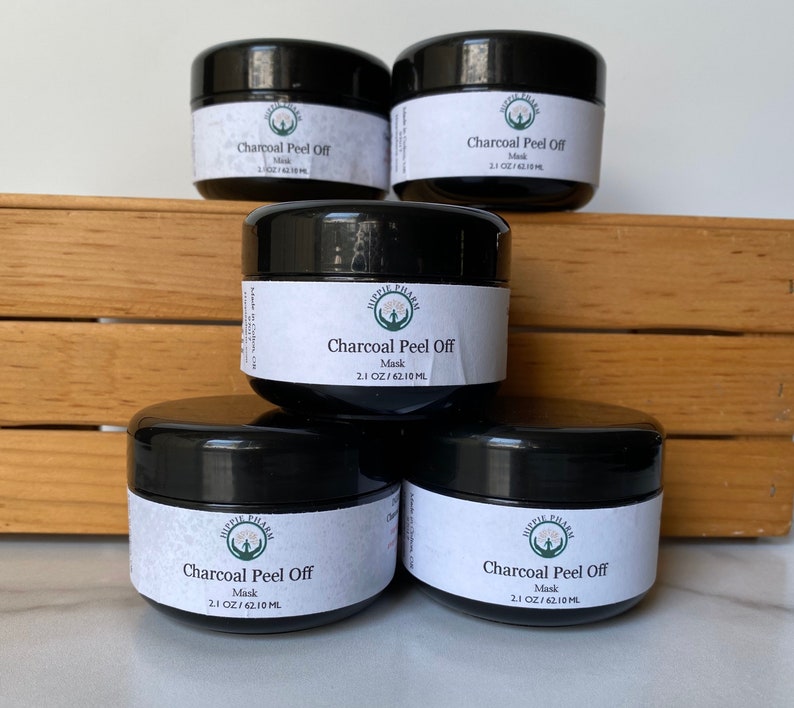 May include: Four black jars of charcoal peel off face mask, each labeled "Hippie Pharm Charcoal Peel Off Mask 2.1 OZ/62.10 ML" on a wooden surface.