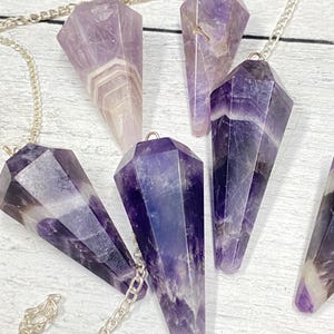 May include: Six amethyst crystal pendulums with silver chains. The pendulums are pointed and have a purple and white color pattern.