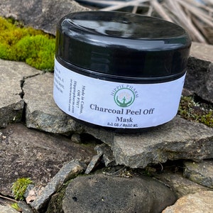 May include: A black jar with a white label that reads "Hippie Pharm" and "Charcoal Peel Off Mask 2.1 oz / 62.10 ml". The jar is sitting on a pile of rocks.