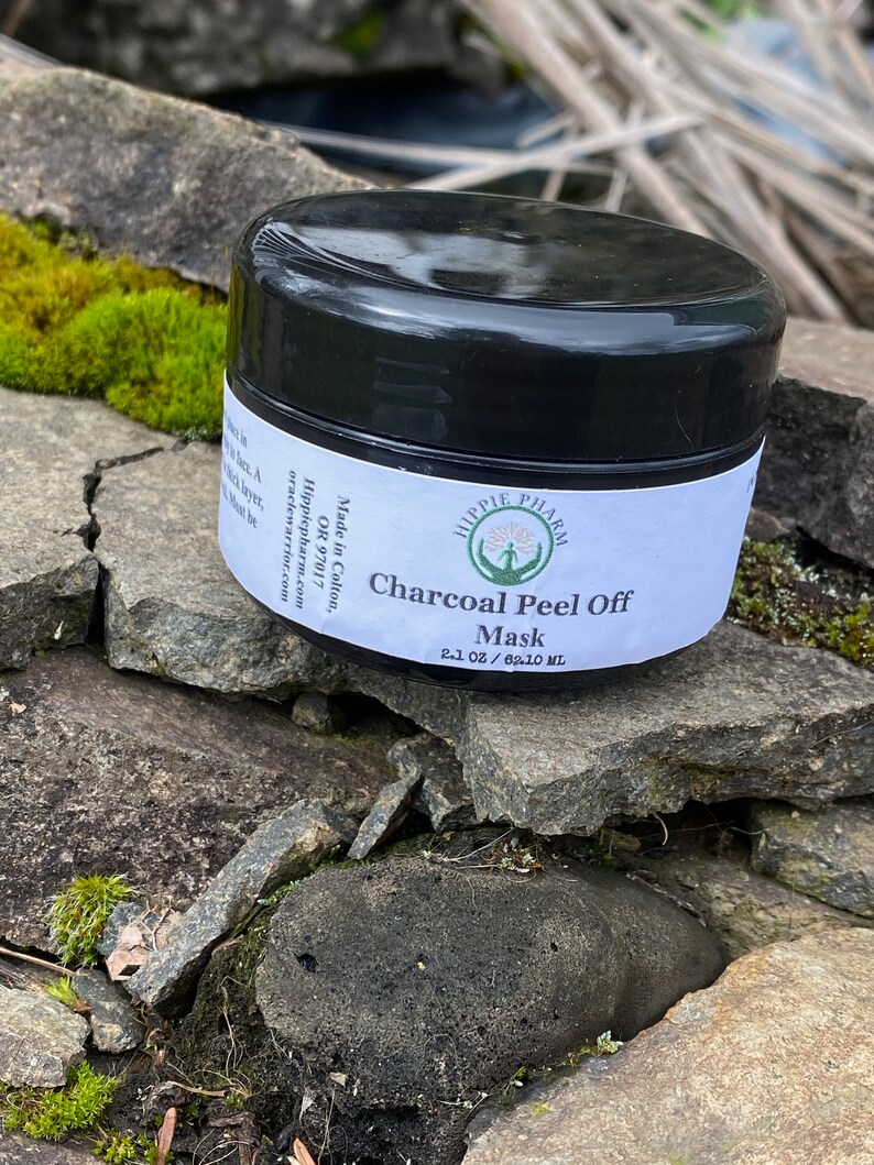 May include: A black jar with a white label that reads "Hippie Pharm" and "Charcoal Peel Off Mask 2.1 oz / 62.10 ml". The jar is sitting on a pile of rocks.
