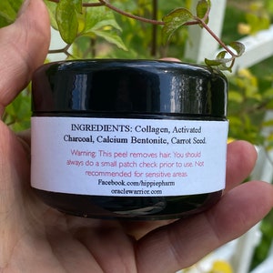 May include: A black jar with a white label that reads "INGREDIENTS: Collagen, Activated Charcoal, Calcium Bentonite, Carrot Seed. Warning: This peel removes hair. You should always do a small patch check prior to use. Not recommended for sensitive areas. Facebook.com/hippiepharm oraclewarrior.com"