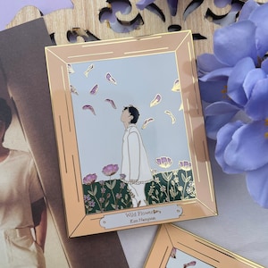 May include: A gold-framed enamel pin featuring a man walking through a field of flowers. The pin has a light blue background with white petals falling from the sky. The text "Wild Flower" and "Kim Namjoon" is printed on the bottom of the pin.