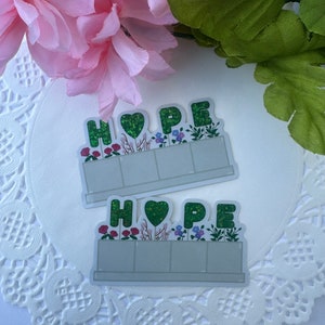 May include: Two stickers with the word "HOPE" in green glitter letters with a green heart in the middle of the word. The letters are surrounded by a green border with flowers and plants. The stickers are on a white lace background.