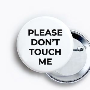 May include: A round white pin-back button featuring the black text PLEASE DON'T TOUCH ME centered on the front. The circular accessory has a smooth surface and a metallic silver pin mechanism visible on the back side of the button design.