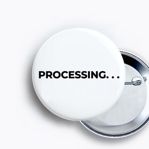 May include: A round white pin-back button featuring the black text "PROCESSING..." printed in a bold sans-serif font. The button has a metallic silver backing with a pin mechanism visible on the right side, showing the construction of the item.