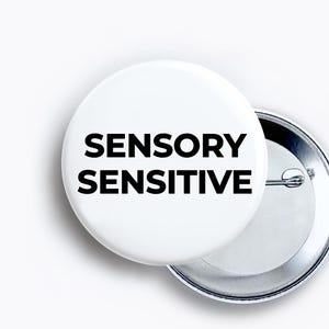May include: A circular pinback button featuring a white background with the text SENSORY SENSITIVE printed in bold black capital letters. The button has a metallic silver backing with a pin mechanism visible on the right side of the object.