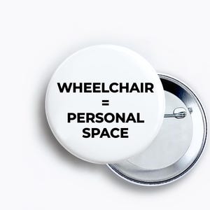 May include: A round white pin-back button featuring the black text WHEELCHAIR = PERSONAL SPACE centered on the front. The button has a metallic silver backing with a pin mechanism visible on the right side, showing the construction of the item.