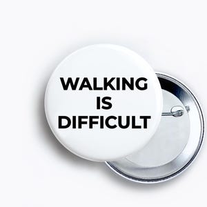 May include: A round white pin-back button featuring the black text WALKING IS DIFFICULT printed in a bold sans-serif font. The circular button has a metallic silver backing with a pin mechanism visible on the right side of the object.
