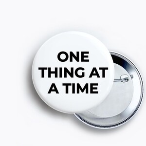 May include: A circular pin-back button with a white background and black text that reads ONE THING AT A TIME. The button features a metallic silver backing with a pin mechanism visible on the right side, showing the construction of the accessory.