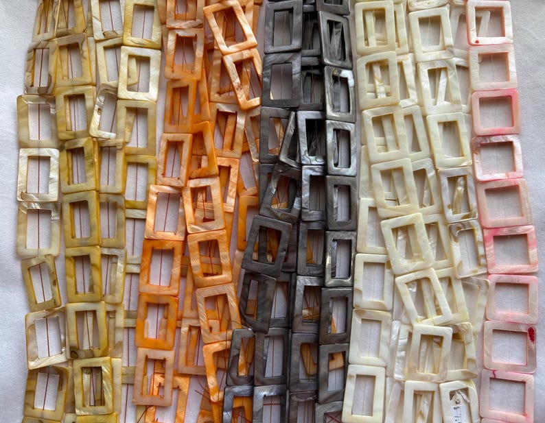 Op de afbeelding: Rijen vierkante gesp gespen in verschillende kleuren. De kleuren zijn geel, oranje, grijs, wit en roze. De gespen zijn gemaakt van een natuurlijk materiaal en hebben een glanzende afwerking. De gespen zijn gerangschikt in verticale rijen.