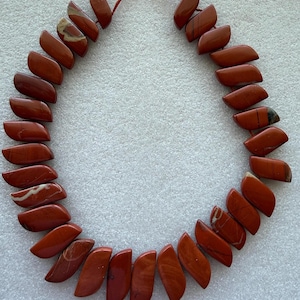 May include: A necklace composed of polished, reddish-brown jasper stones. Each stone is a unique shape, strung together on a red cord. The stones have natural patterns and variations in color, creating a striking visual appeal.
